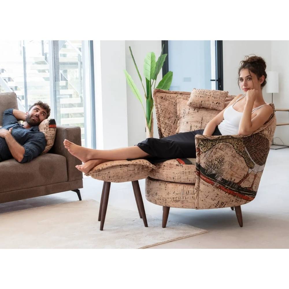 A woman relaxes in a patterned beige armchair with a matching footrest in a modern living room, while a man lounges on a brown sofa in the background.