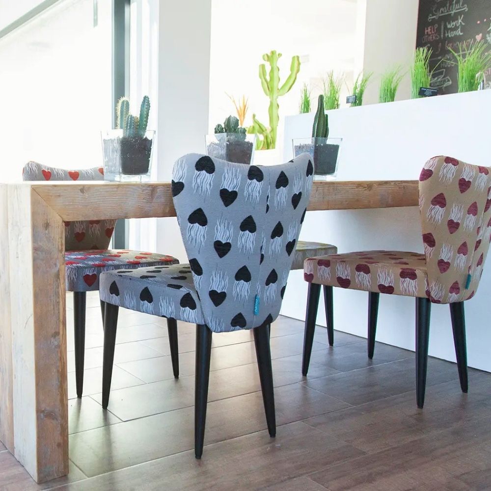 A dining area scene with a wooden table and upholstered chairs. The main visible chair in the foreground has a light gray fabric with black heart patterns, while other chairs show different upholstery colors and patterns.