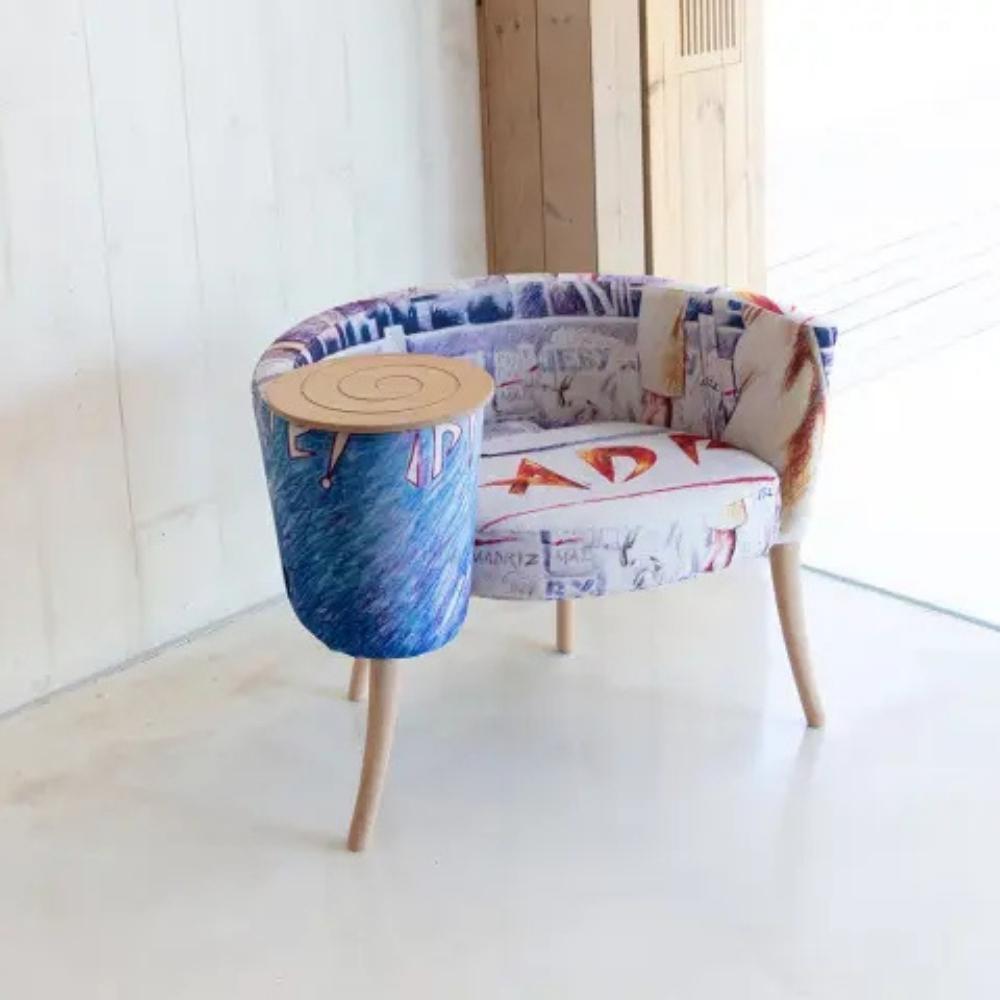 A modern upholstered lounge chair with a cylindrical side table attached to the right side. The chair features a curved backrest and seat cushion upholstered in a multicolor abstract patterned fabric, supported by light natural wood legs.