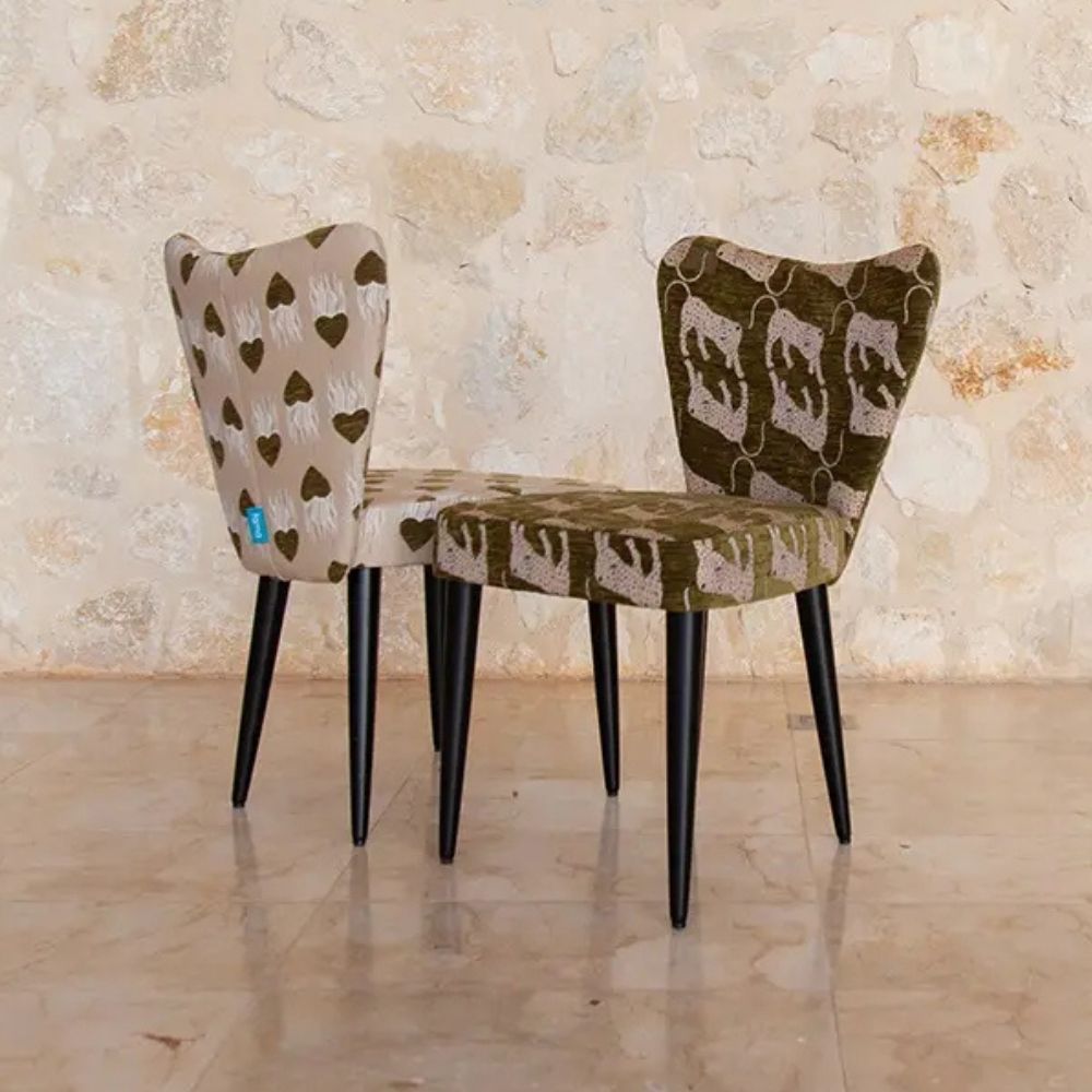 Two upholstered dining chairs with black tapered legs, shown in front of a textured stone wall and tiled floor. One chair has a beige backrest with dark olive heart motifs and a matching seat, while the other has a dark olive patterned backrest and a beige patterned seat.
