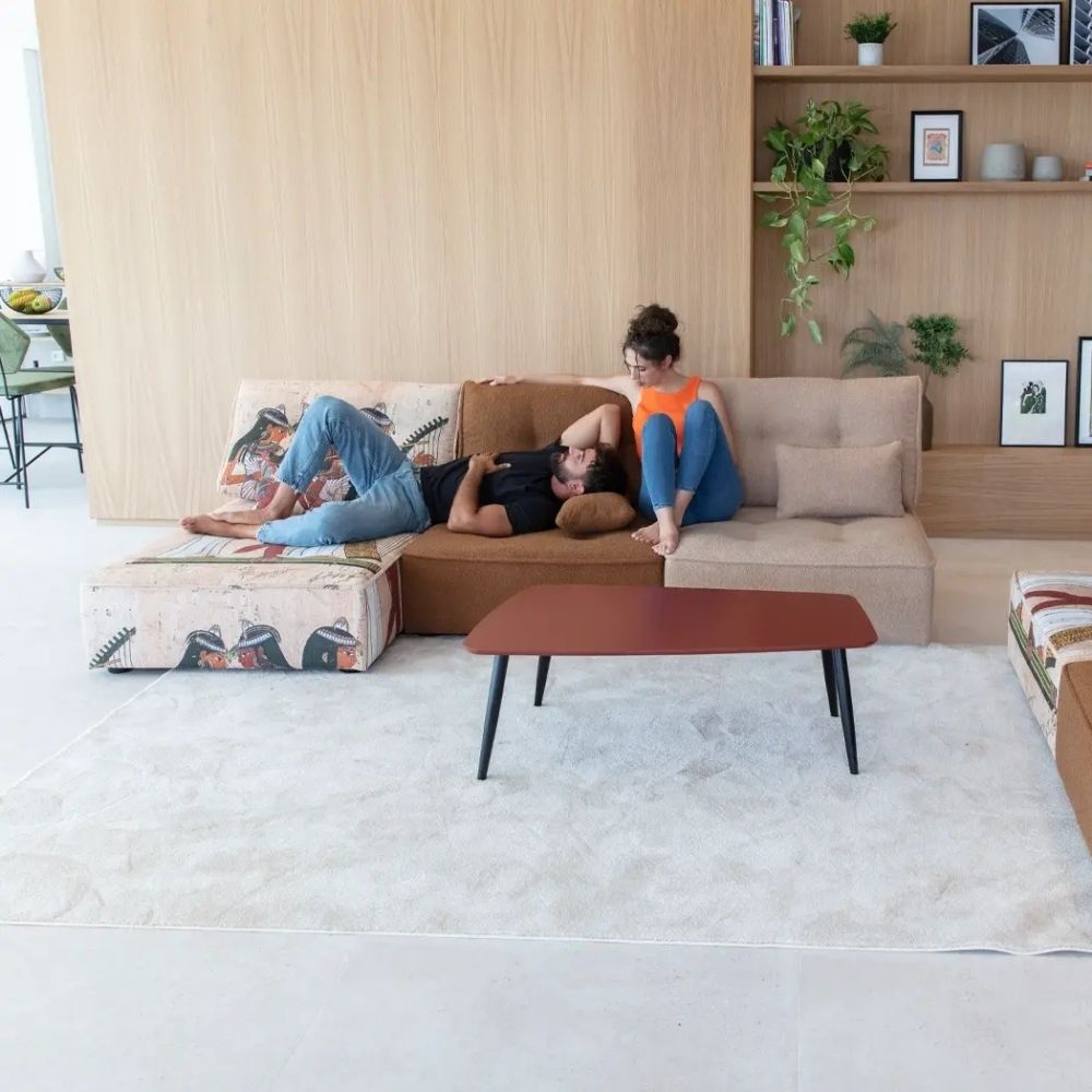 A modern living room scene with two people relaxing on a sectional sofa. One person lies on the left on a patterned ottoman/sofa section, and the other sits on the right on a beige sofa section.