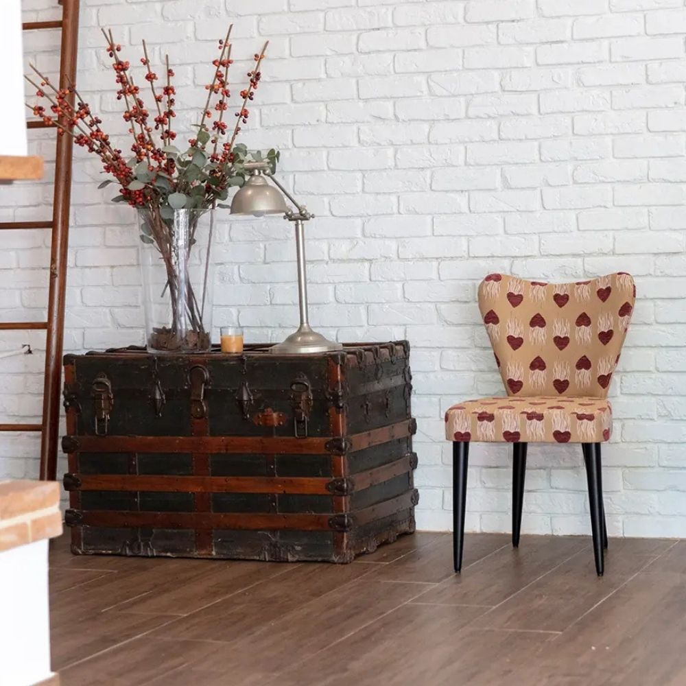 A vintage-style upholstered chair with a beige fabric and red heart pattern, positioned next to a large dark wooden trunk against a white brick wall.