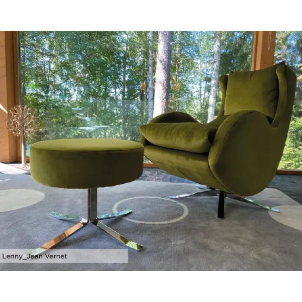 An olive green velvet lounge chair with a matching round footstool, set in a modern room with a large window overlooking a forest. The chair has a curved backrest and arm, and the footstool has a chrome star base.