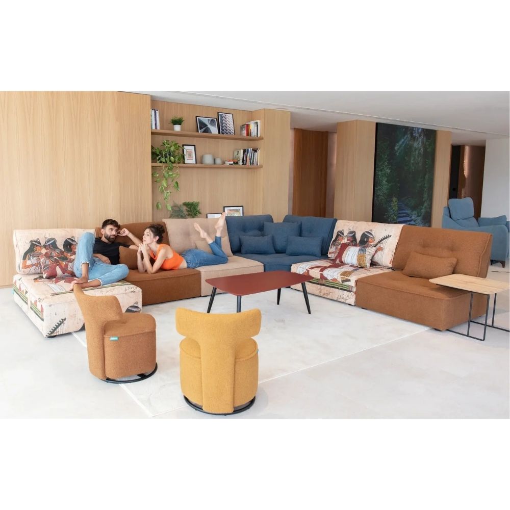 A modern living room set featuring a brown sectional sofa with patterned throw pillows, a blue sectional sofa, two mustard yellow armchairs, and a small wooden coffee table, with two people lounging on the brown sofa in a bright, contemporary interior.