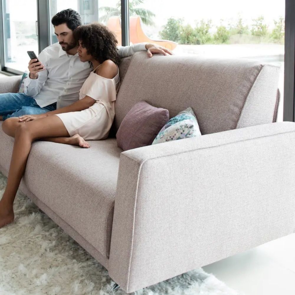 A man and a woman sit closely together on a light beige upholstered sofa in a bright modern room, with the man holding a phone and large windows behind them.