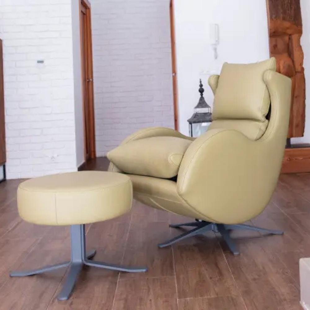 A light beige faux leather recliner chair with a matching round footstool, set on a dark wood floor in a room with white brick walls.