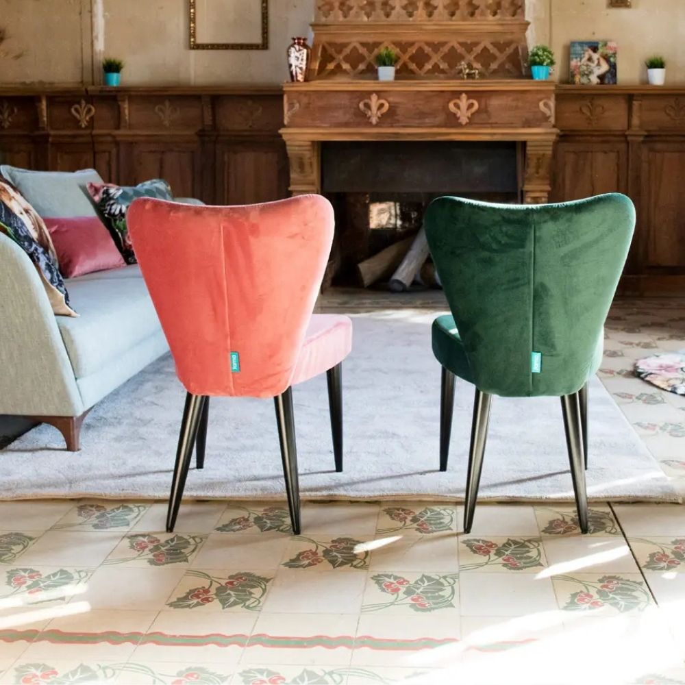 Two velvet accent chairs in coral pink and emerald green, shown from the back in a styled living room setting with a fireplace and patterned rug.