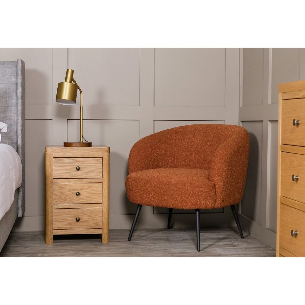 An orange-brown upholstered armchair with black metal legs, shown in a styled bedroom setting next to a light wood three-drawer nightstand and a brass table lamp.