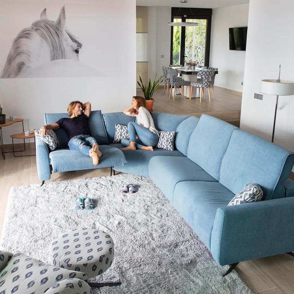 A light blue sectional sofa in a modern living room, with two people lounging on the sofa. The room features a large white wall with a black and white horse print, a white shag rug, and a dining area in the background.