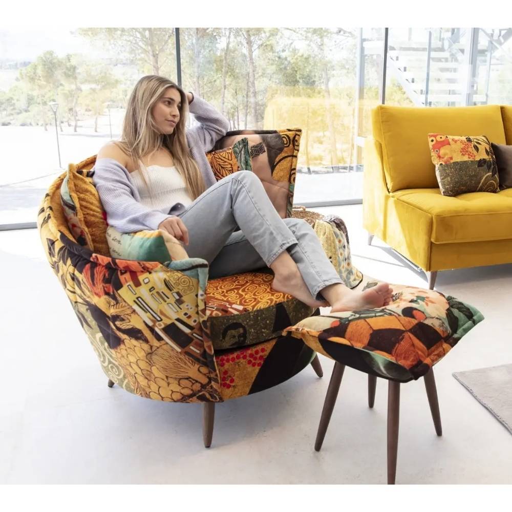 A woman sits in a round upholstered armchair with a matching footrest, both covered in a colorful abstract patterned fabric. The chair and stool are placed in a bright modern room with large windows, and a yellow sofa is visible in the background.
