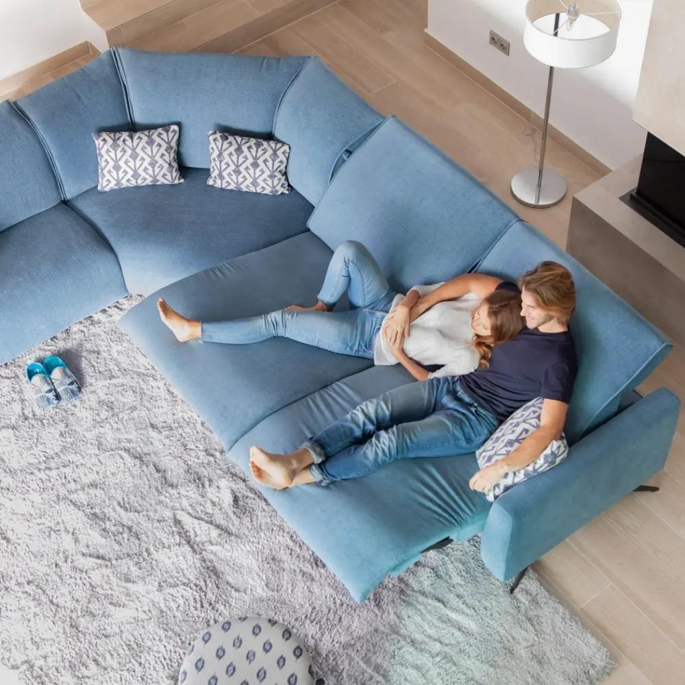 A blue sectional sofa in a modern living room, with a couple lying comfortably on the couch. The scene is captured from an overhead angle, showing the sofa's L-shape design, patterned throw pillows, and a light gray shag rug.