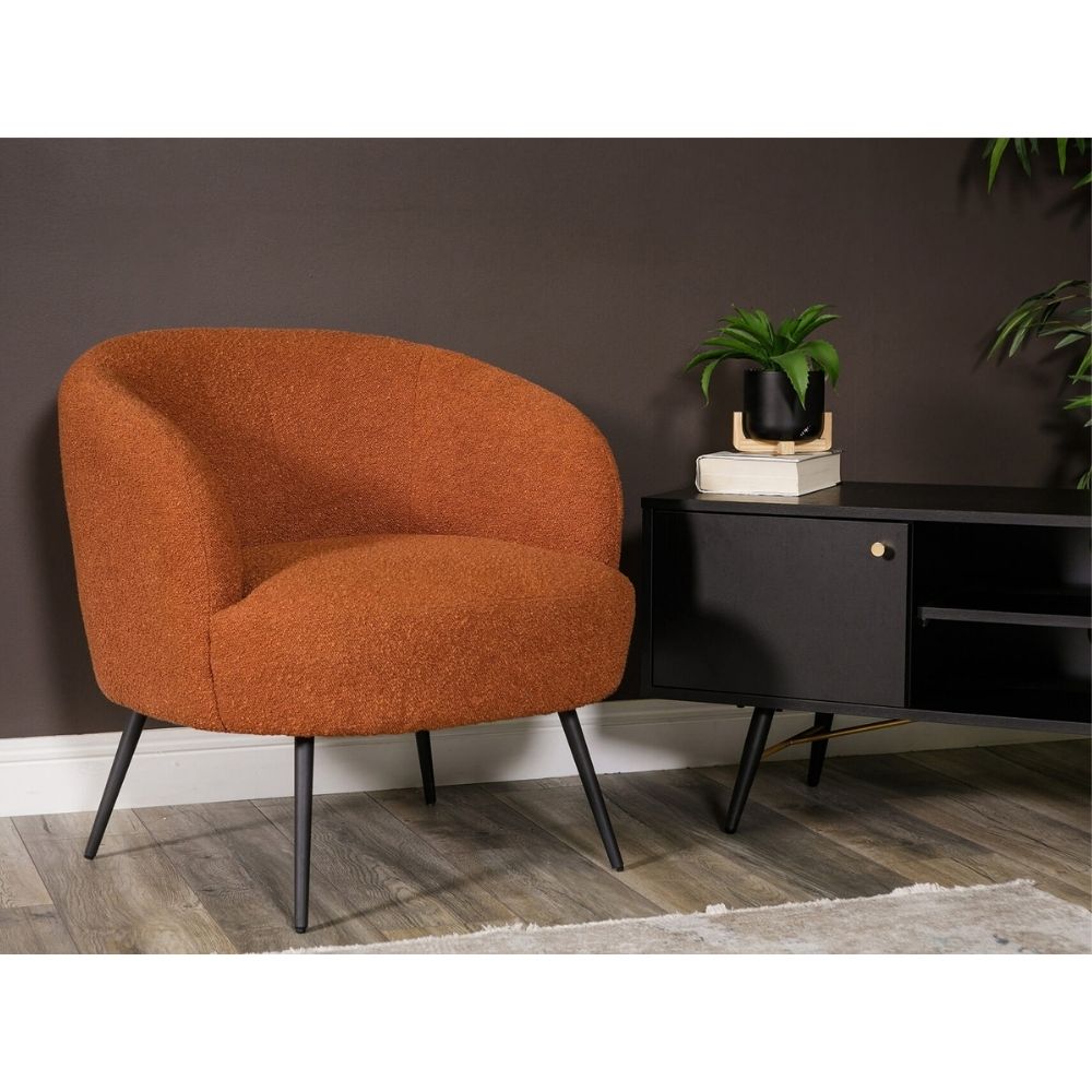 An orange-brown textured barrel chair with black metal legs, positioned next to a black sideboard in a modern room setting with a dark brown wall and wood-look flooring.