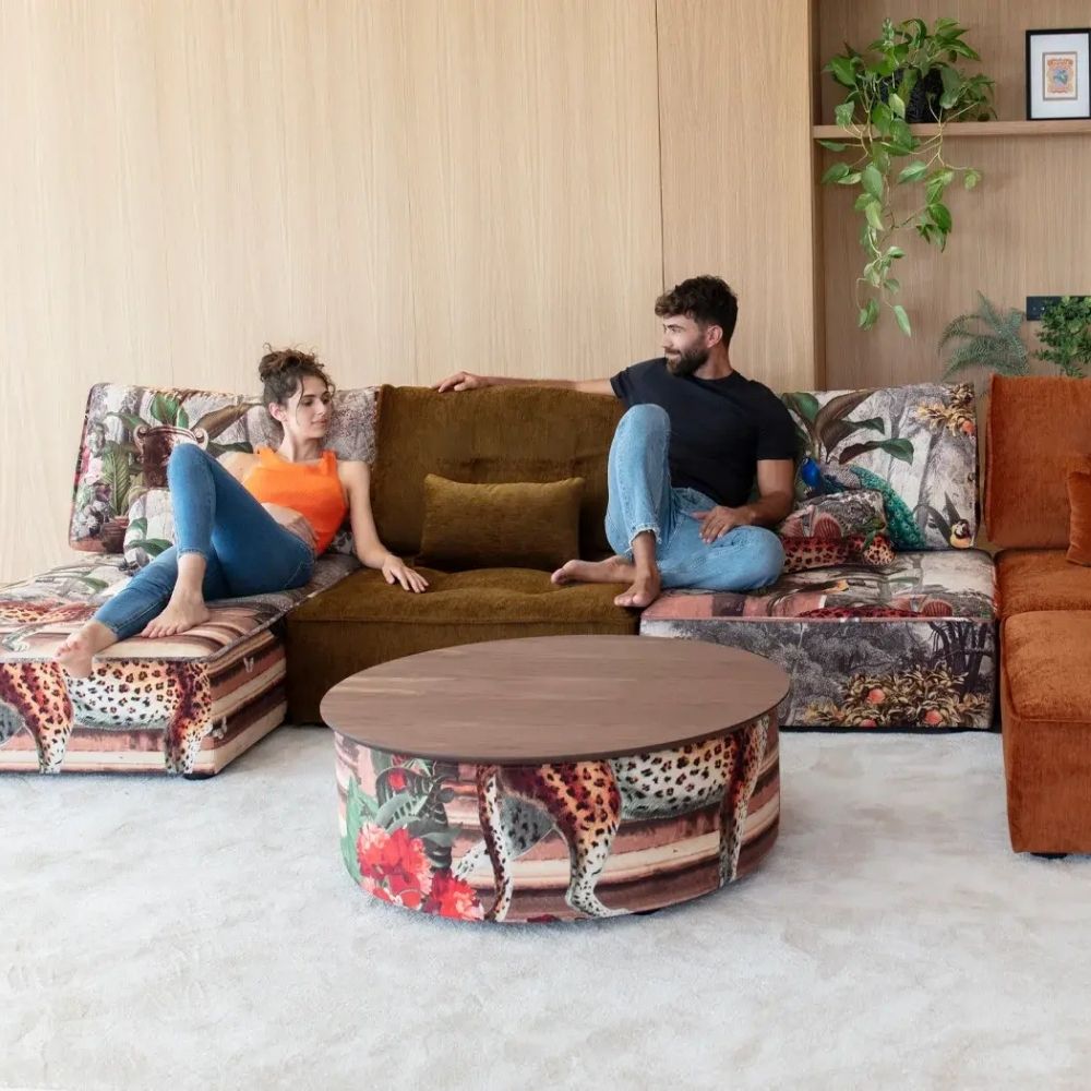 A man and a woman relax on a large sectional sofa in a modern living room. The sectional features a mix of colorful animal-print upholstery and a central olive green velvet section, with a matching round coffee table in front.