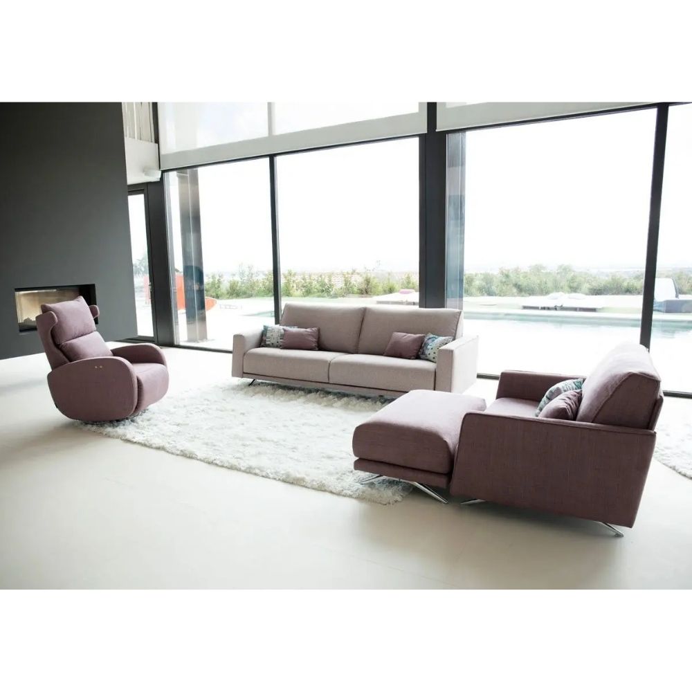 A modern living room furniture set featuring a mauve-pink armchair, matching ottoman, and a light beige sofa with pillows, arranged on a white shag rug in front of large floor-to-ceiling windows.