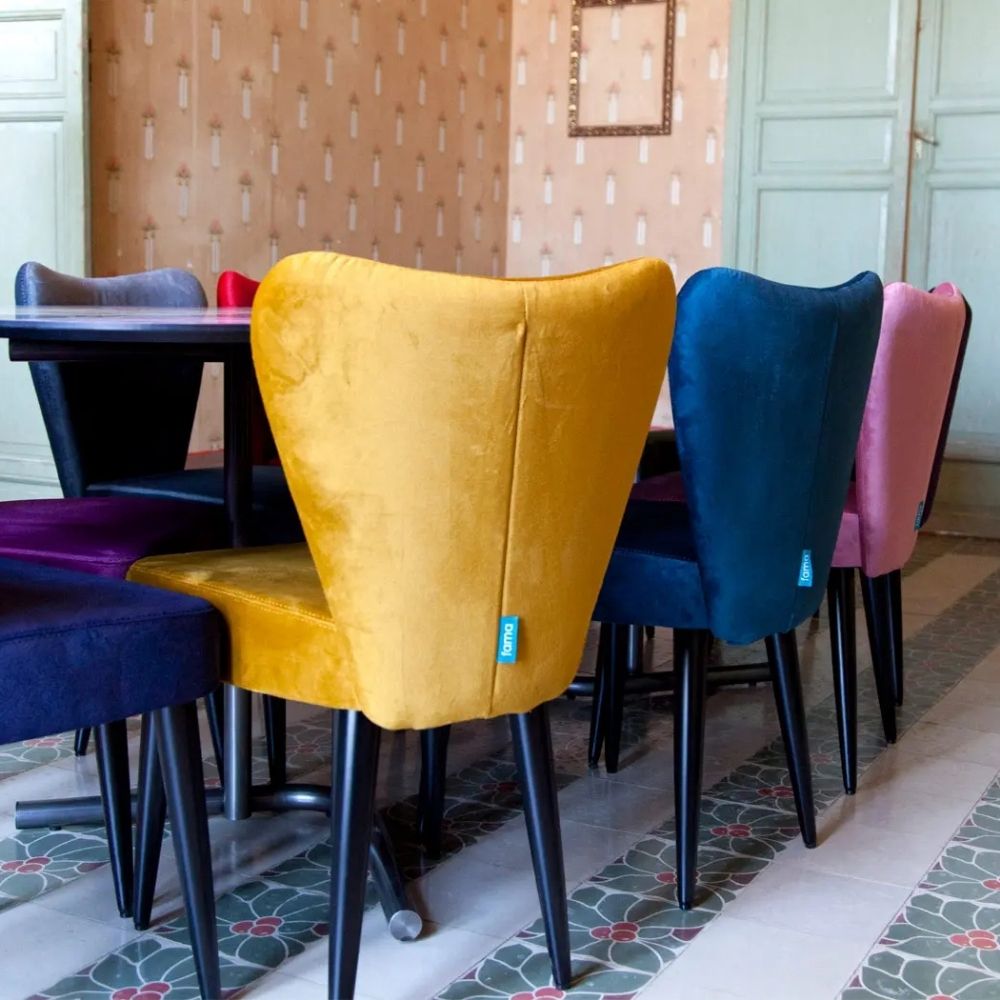 A set of velvet dining chairs in mustard yellow, teal blue, pink, and purple, arranged around a dark wooden dining table in a dining room with patterned wallpaper and tiled floors.