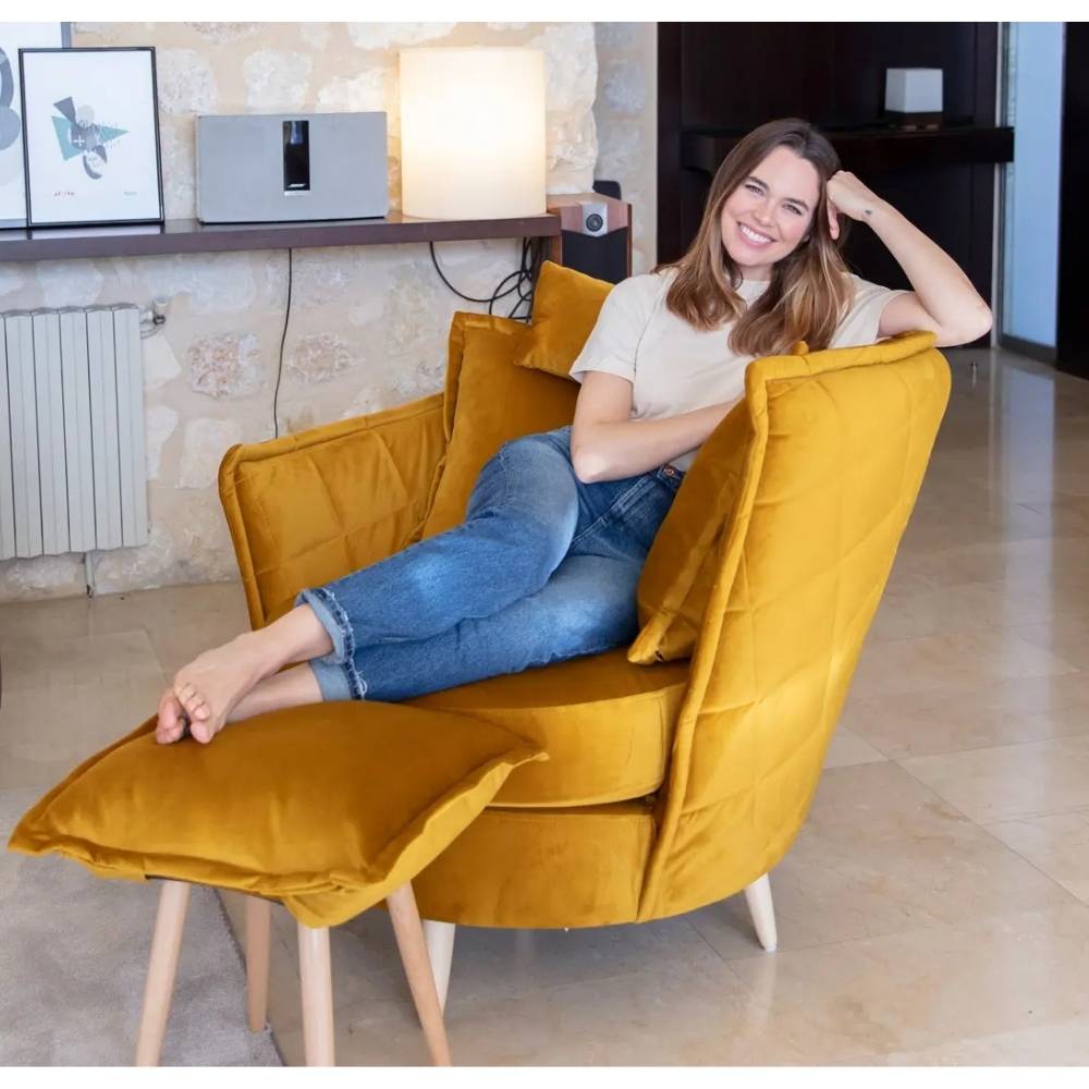 A woman reclining in a mustard yellow velvet armchair with a matching footrest in a modern living room setting.