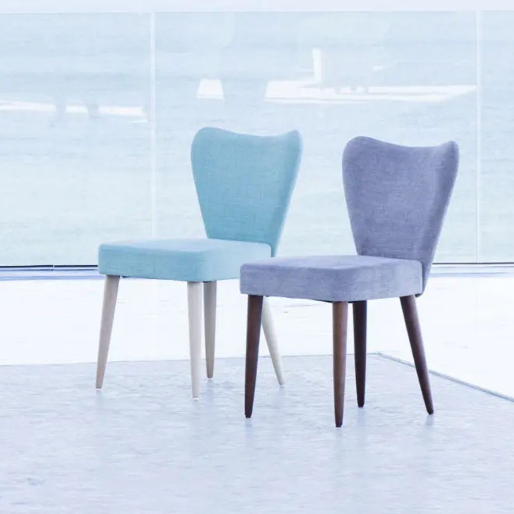 Two modern upholstered dining chairs with winged backs are shown in a bright, minimalist interior. One chair is light aqua blue with pale wood legs, and the other is lavender-gray with dark wood legs.