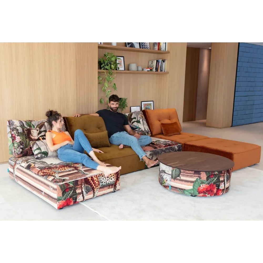 A modern sectional sofa set with a brown leather-like seat, orange upholstered side sections, and patterned floral pillows and floor cushions, shown in a living room with two people reclining on the sofa. A round coffee table with matching floral upholstery sits in front.