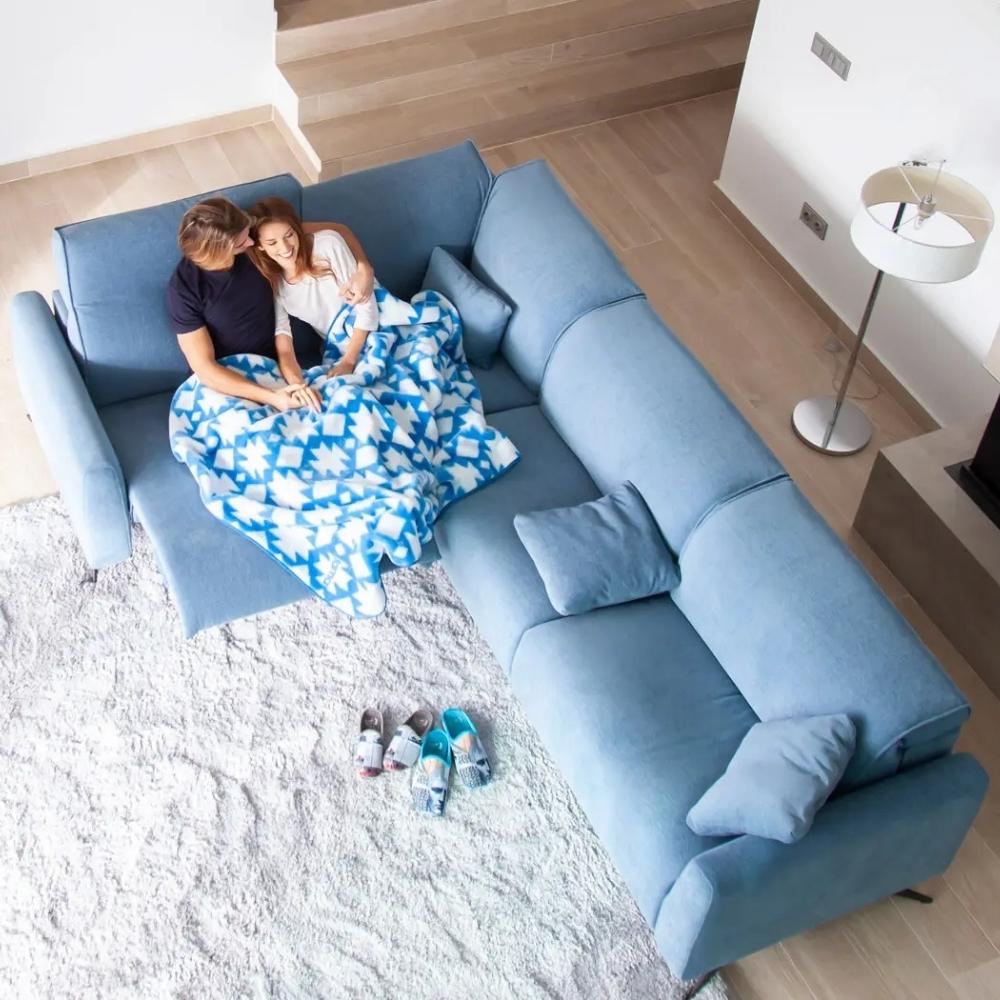 A high-angle view of a modern light blue sectional sofa in a bright living room, with a couple sitting underneath a blue and white patterned blanket on the sofa.