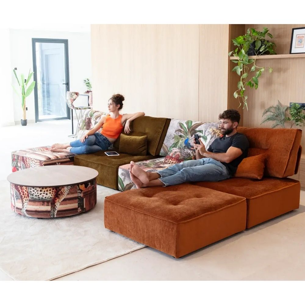 A modern living room scene showing a large rust-orange modular sectional sofa with two people lounging on it. One person is reclining on the left side of the sofa, and the other is lying down on the chaise section on the right.