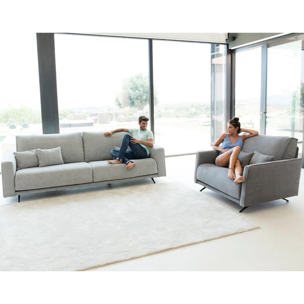 A man and a woman relax on two light gray upholstered sofas in a bright, modern living room with large floor-to-ceiling windows and a white rug.