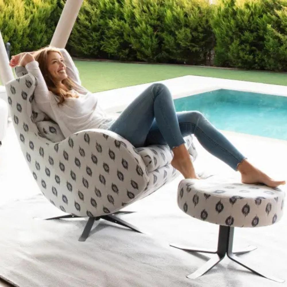 A woman reclines in a modern swivel lounge chair with a matching footstool beside a swimming pool. The chair and footstool have a light beige fabric with a repeating dark gray abstract leaf-like pattern, and the chairs have black metal swivel bases.