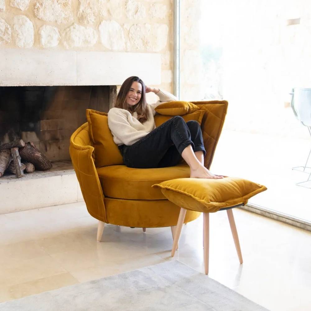 A woman relaxes in a mustard yellow velvet armchair with a matching footrest, set in a bright room with a stone fireplace and large windows.