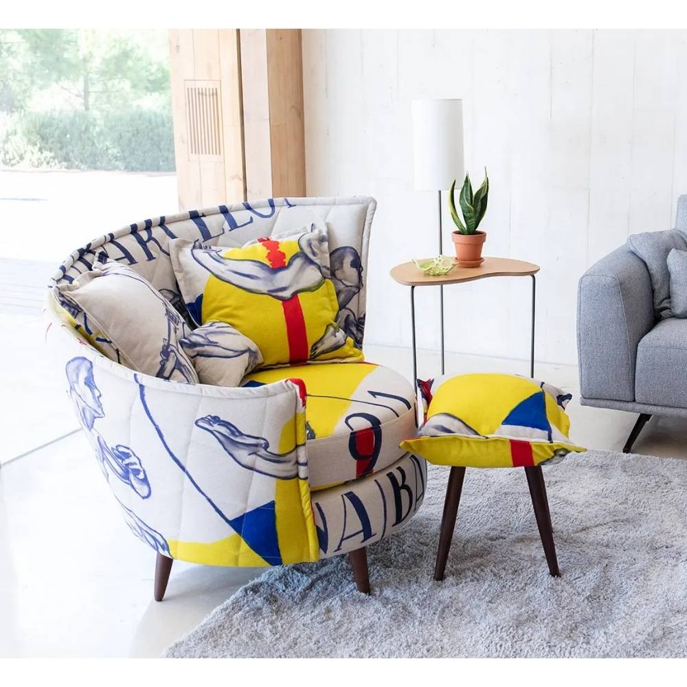 A modern curved armchair with a matching footstool and throw pillows, upholstered in a colorful abstract print featuring yellow, blue, red, and white, styled in a bright living room setting.