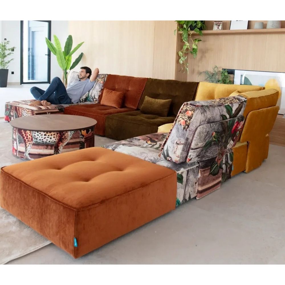 A modern sectional sofa in a bright living room, featuring rust orange velvet, mustard yellow velvet, and a central floral camouflage print fabric. A man is lounging on the sofa next to a round coffee table.