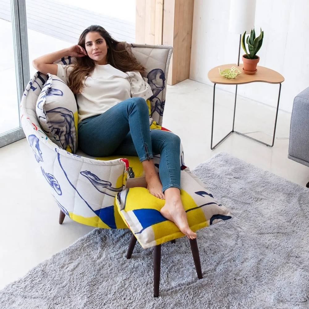 A woman reclines in a modern upholstered armchair with a matching footrest in a bright, contemporary room. The chair features a white base with blue abstract line art and bold yellow and blue geometric blocks on the seat and backrest, supported by dark wooden legs.