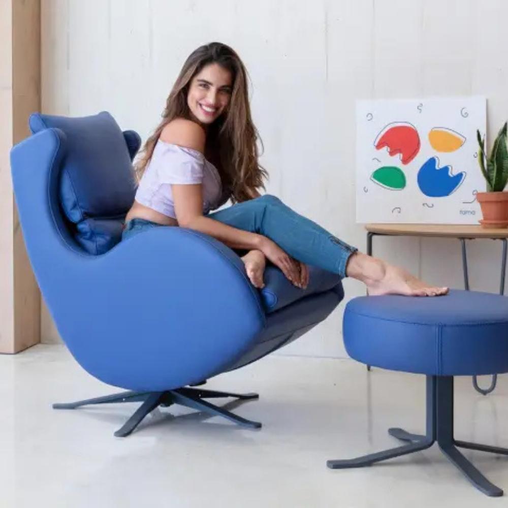 A woman sits barefoot in a blue swivel lounge chair with a matching blue footstool in a bright indoor room. The chair has a rounded shell design and black star base, with a small side table and colorful wall art in the background.