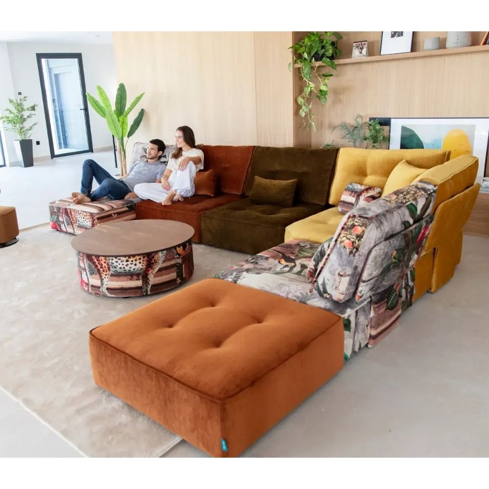 A modern sectional sofa set in a bright, contemporary living room. The set includes a rust-orange tufted ottoman in the foreground, a dark brown velvet section, a mustard yellow armchair, and a rust-brown leather section.