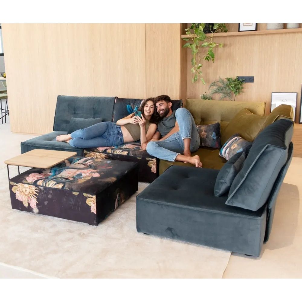 A modern sectional sofa set with a dark teal velvet chaise lounge, a matching tufted ottoman with a floral print, and a yellow-green accent sofa, shown with a couple lounging on the sofa in a bright, contemporary living room.