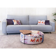 A light gray fabric sofa with two patterned throw pillows, positioned in a living room setting with a round patterned ottoman and a small wooden side table in front of it.