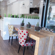 A rustic wooden dining table in a bright modern kitchen, surrounded by patterned dining chairs including one with red hearts on a white background and another with yellow stars on a white background. Small potted cacti sit on the table.