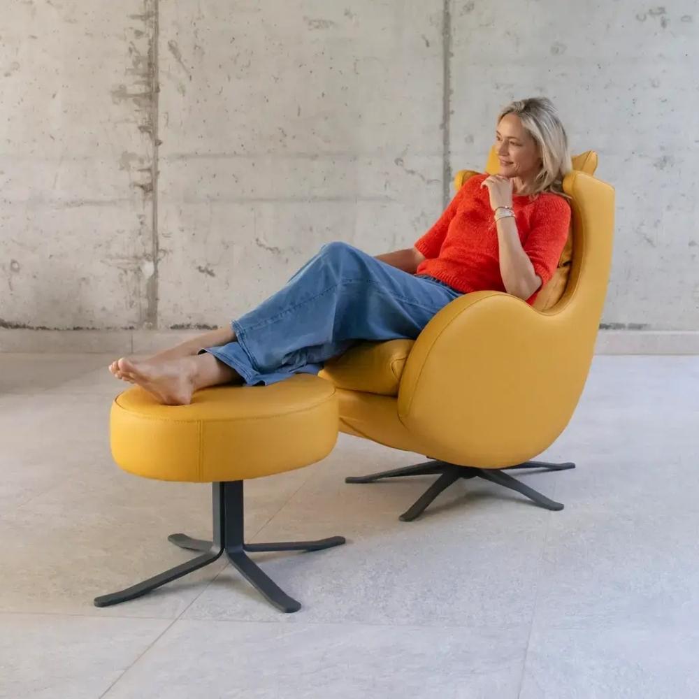 A woman reclining in a modern mustard yellow lounge chair with an attached matching footstool, set against a concrete wall and floor.