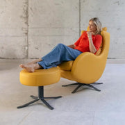 A woman reclining in a modern mustard yellow lounge chair with an attached matching footstool, set against a concrete wall and floor.