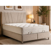A beige upholstered ottoman divan bed frame with a matching tufted headboard and a white quilted mattress, shown in a styled bedroom setting with a lamp, framed wall art, and a potted plant.