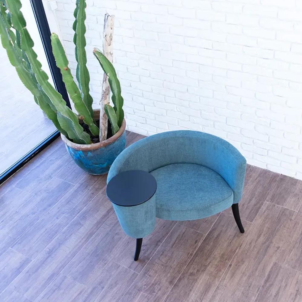 A modern curved lounge chair in light blue fabric with a matching cylindrical side table, placed on a wood-look floor against a white brick wall next to a large potted cactus.