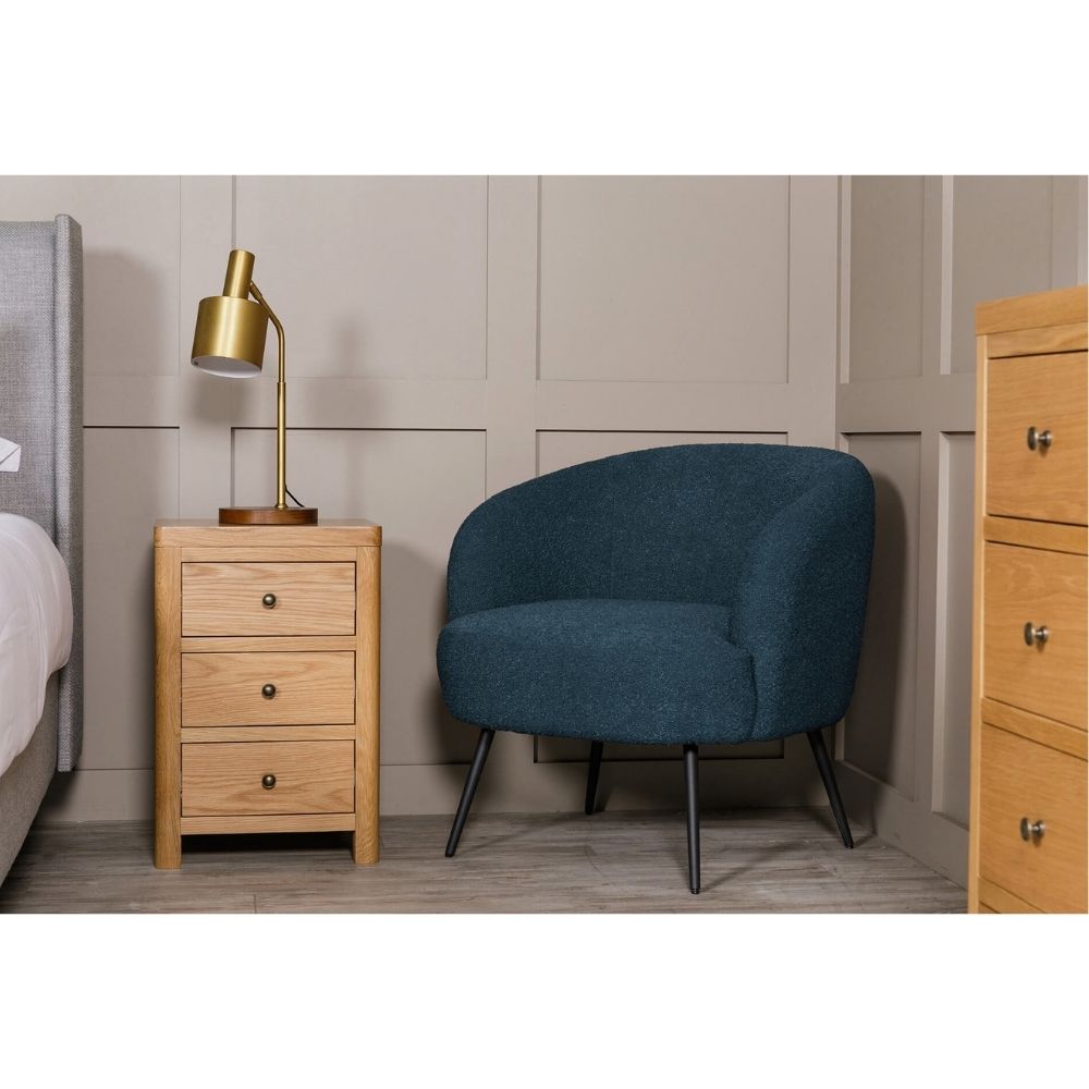 A modern navy blue upholstered barrel chair with black metal legs, styled in a bedroom setting next to a light oak three-drawer nightstand with a gold desk lamp.