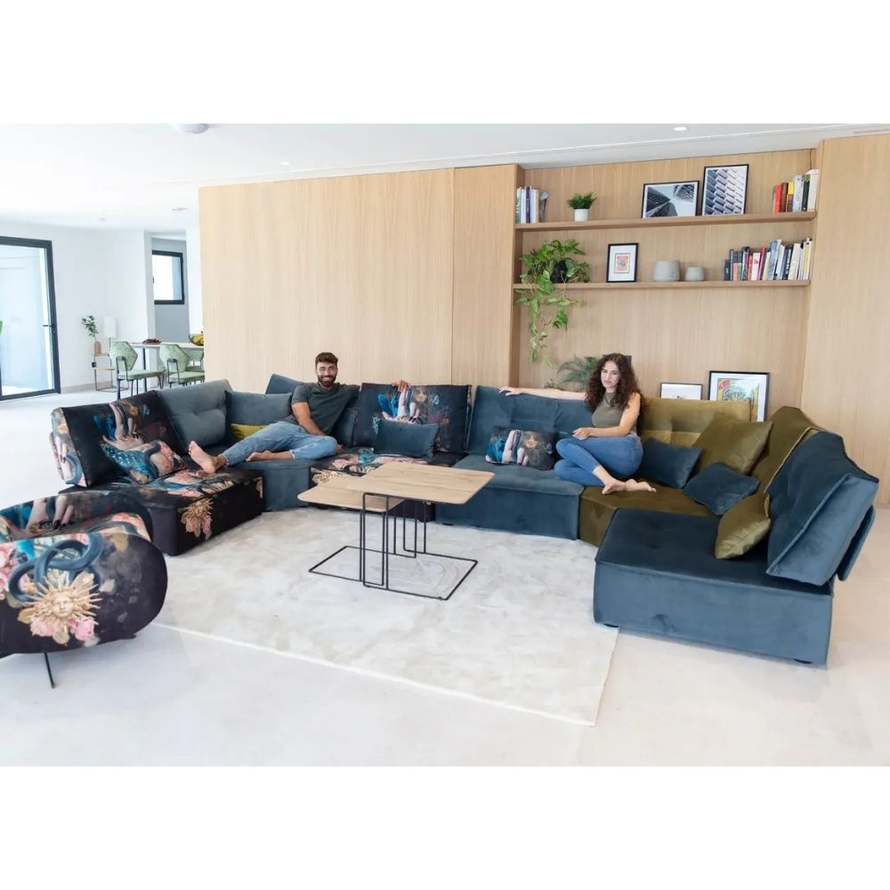 A modern sectional sofa set in a bright, contemporary living room. The set includes a large U-shaped sectional in slate blue fabric with olive green and mustard yellow accent pillows, a matching floral-patterned armchair, and a small wooden coffee table on a white rug.