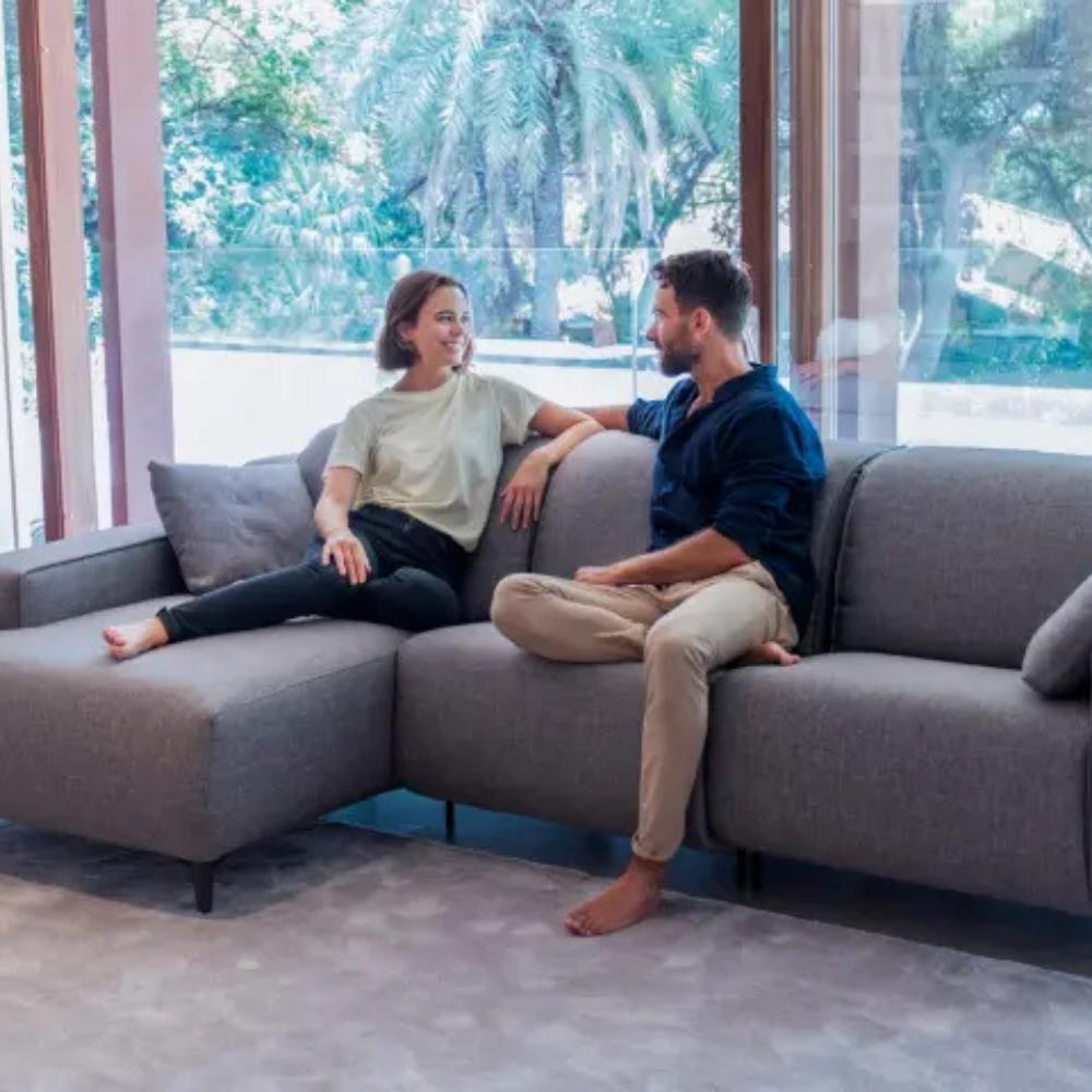 A man and a woman sitting on a modern gray sectional sofa in a bright room with large windows and outdoor greenery visible behind them.