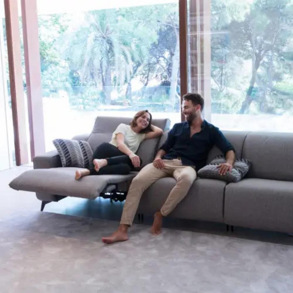 A man and a woman relax on a modern gray sectional sofa with an extended chaise lounge in a bright, contemporary living room with large windows overlooking green trees.