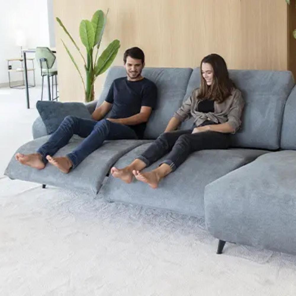 A man and a woman sit barefoot on a large light gray sectional sofa in a bright, modern living room with a wood-paneled wall and a potted plant.