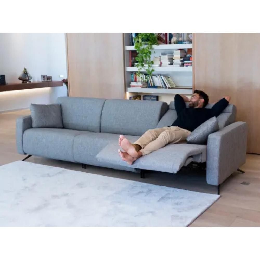 A man relaxes on a light gray upholstered reclining sofa in a modern living room with light wood flooring and built-in shelves.