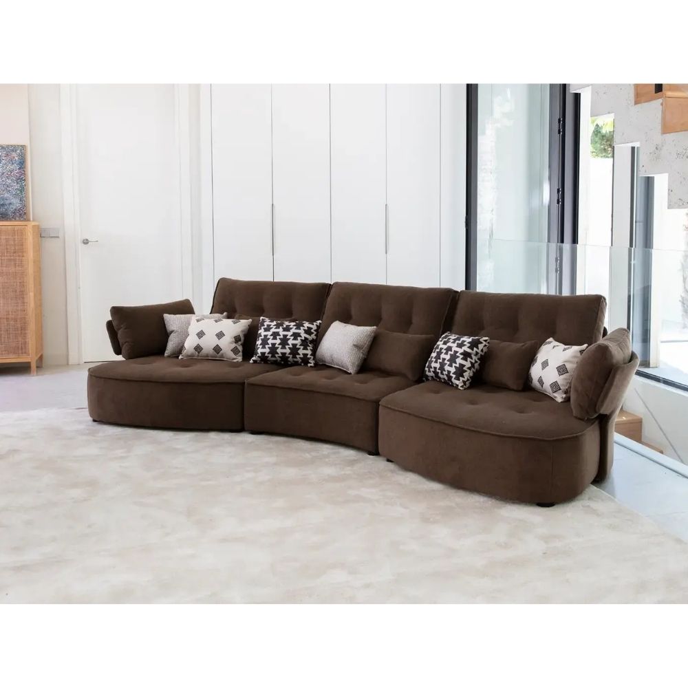 A curved brown fabric sectional sofa with tufted back cushions, styled with several throw pillows, set in a bright, modern living room on a light-colored rug.