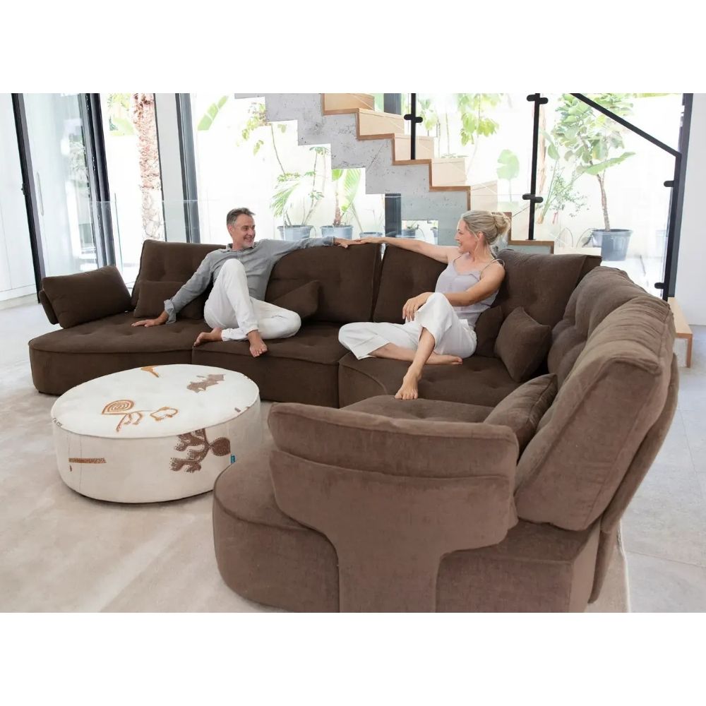 A large brown sectional sofa with two people sitting on it in a bright modern living room, alongside a round white ottoman with a floral pattern.