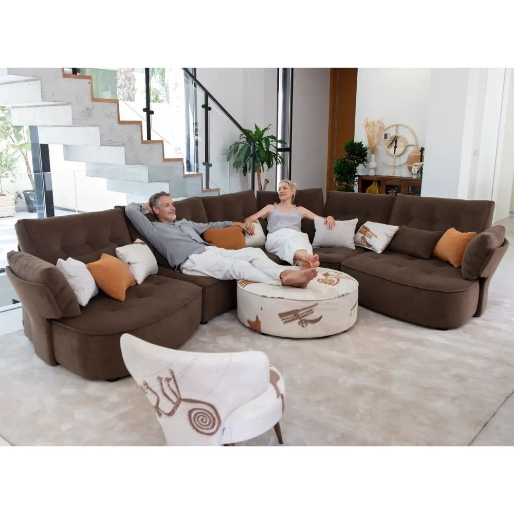A man and a woman relax on a large U-shaped brown sectional sofa in a bright modern living room, with white and orange accent pillows, a round ottoman, and a staircase in the background.