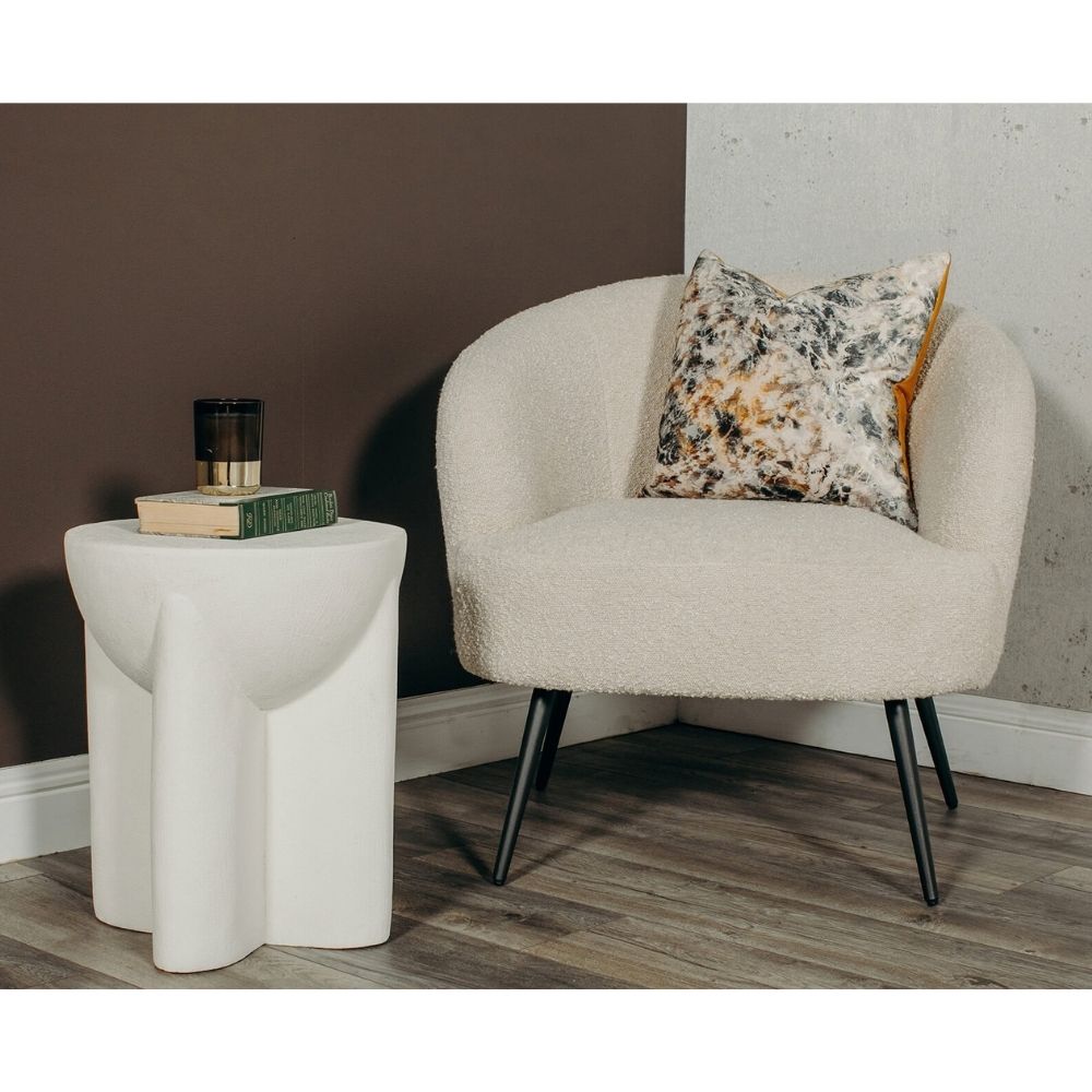 A cream bouclé armchair with black tapered legs sits beside a sculptural white side table in a styled room corner. A patterned throw pillow rests on the chair, and the background features a dark brown wall and a light textured wall.