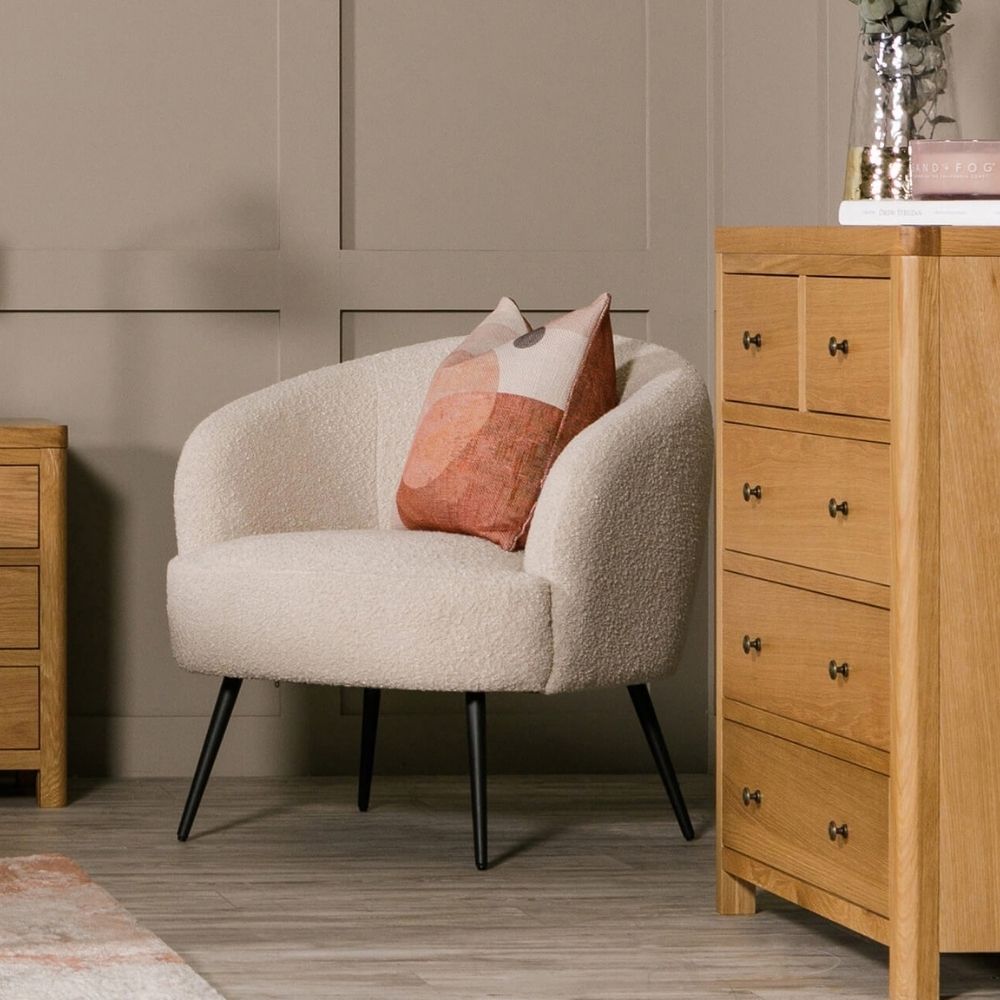 A cream bouclé armchair with black tapered legs, styled in a room with a geometric pink and beige accent pillow, a light oak 5-drawer chest, and a matching side table against a taupe paneled wall.